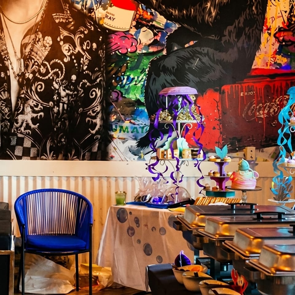 Colorful buffet setup with cupcakes, cakes, and chafing dishes against a vibrant mural.