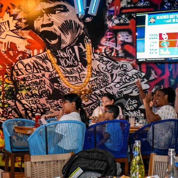 Restaurant patrons watch a sports game on a large screen, with vibrant graffiti art on the wall.