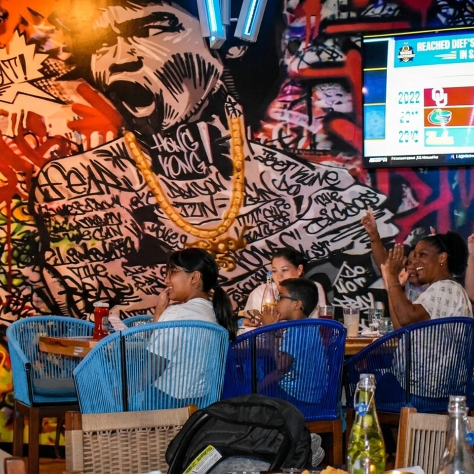 Restaurant patrons watch a sports game on a large screen, with vibrant graffiti art on the wall.