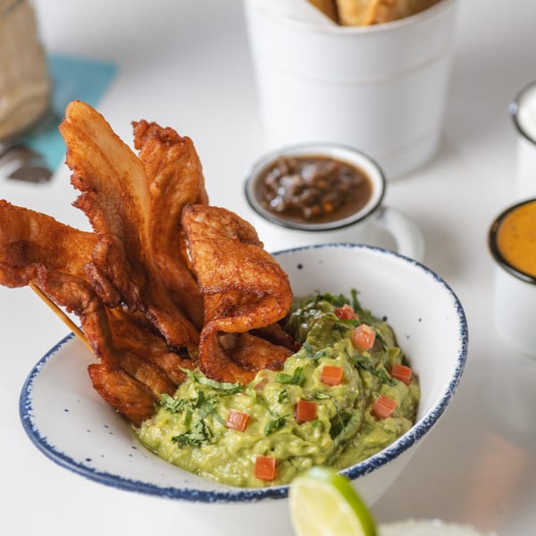 Guacamole with fried pork belly skewers, salsa, and taquitos, with a margarita in the foreground.