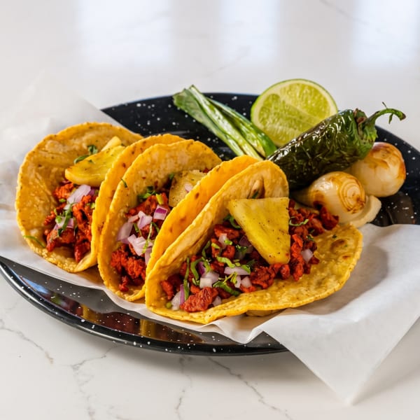 Three al pastor tacos with pineapple, onion, cilantro, lime, grilled pepper, and onions on a plate.