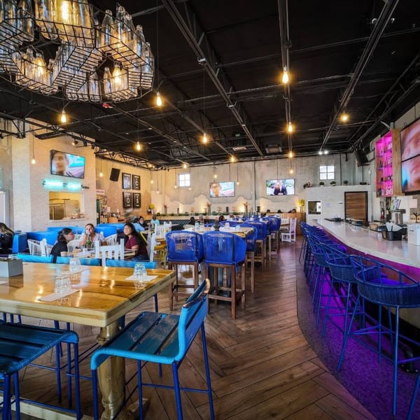 Spacious restaurant interior with blue chairs, wooden tables, a long bar, and people dining.