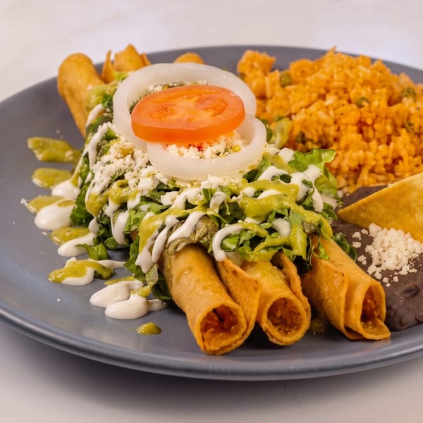 Four golden taquitos topped with lettuce, cheese, tomato, onion, and sauces, served with rice and beans.