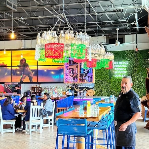 A restaurant interior with a man in an apron, colorful bottle chandeliers, and patrons at tables.