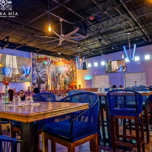 Morra Mia restaurant interior with blue chairs, wooden tables, vibrant wall art, and multiple TV screens.