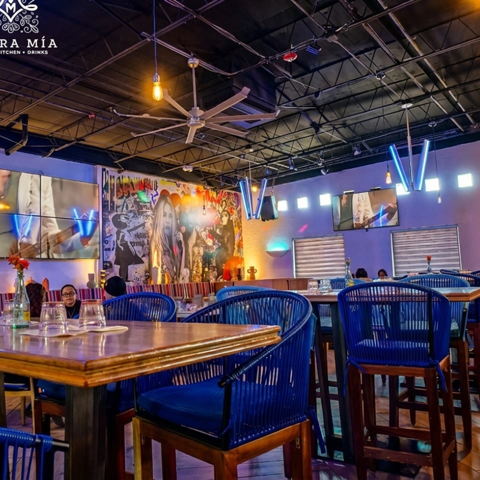 Morra Mia restaurant interior with blue chairs, wooden tables, vibrant wall art, and multiple TV screens.