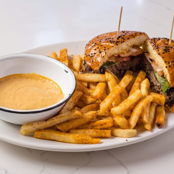 A burger cut in half with fries and a side of dipping sauce on a white plate.