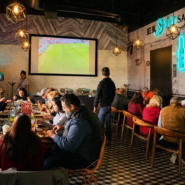 Diners enjoying meals at long tables in a restaurant with a large screen showing a soccer game.